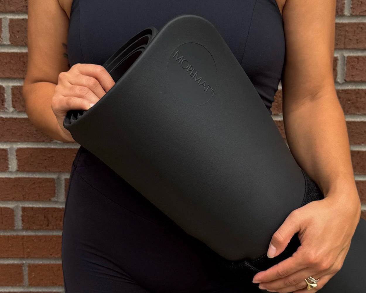 Person holding a black exercise mat with 'MOREMAT' branding against a brick wall.